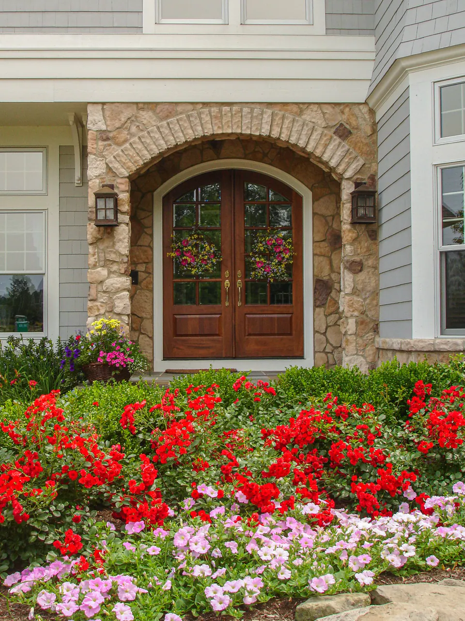 Stone entry arch with double doors, red and pink seasonal flowers, lawn, and brick edging